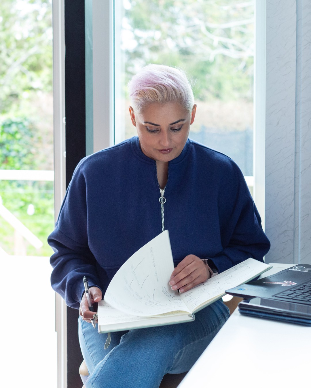A woman writing in a notebook by a window, a laptop beside her